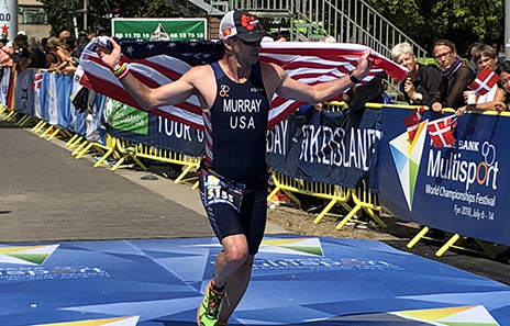 Ian Murray running across the finish line of an Ironman with a US flag around his shoulders